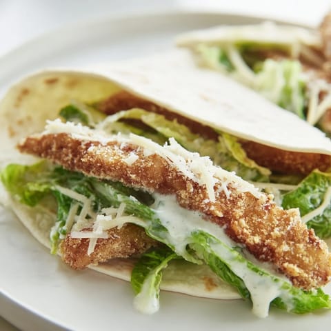 A close-up of a halved Crispy Chicken Caesar Wrap, revealing layers of crunchy romaine, grated Parmesan, and juicy fried chicken coated in creamy Caesar dressing.