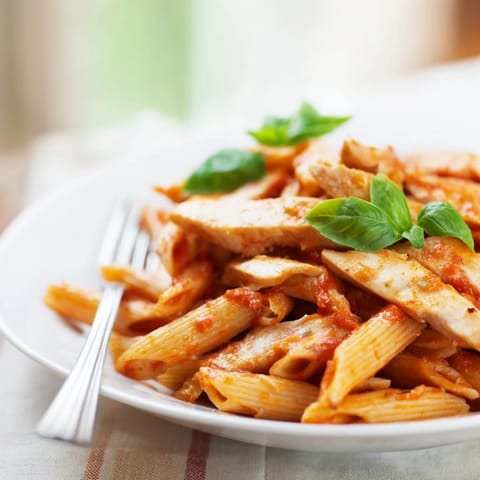 Penne pasta tossed with savory, juicy chicken and a bold red sauce, garnished with fresh basil.
